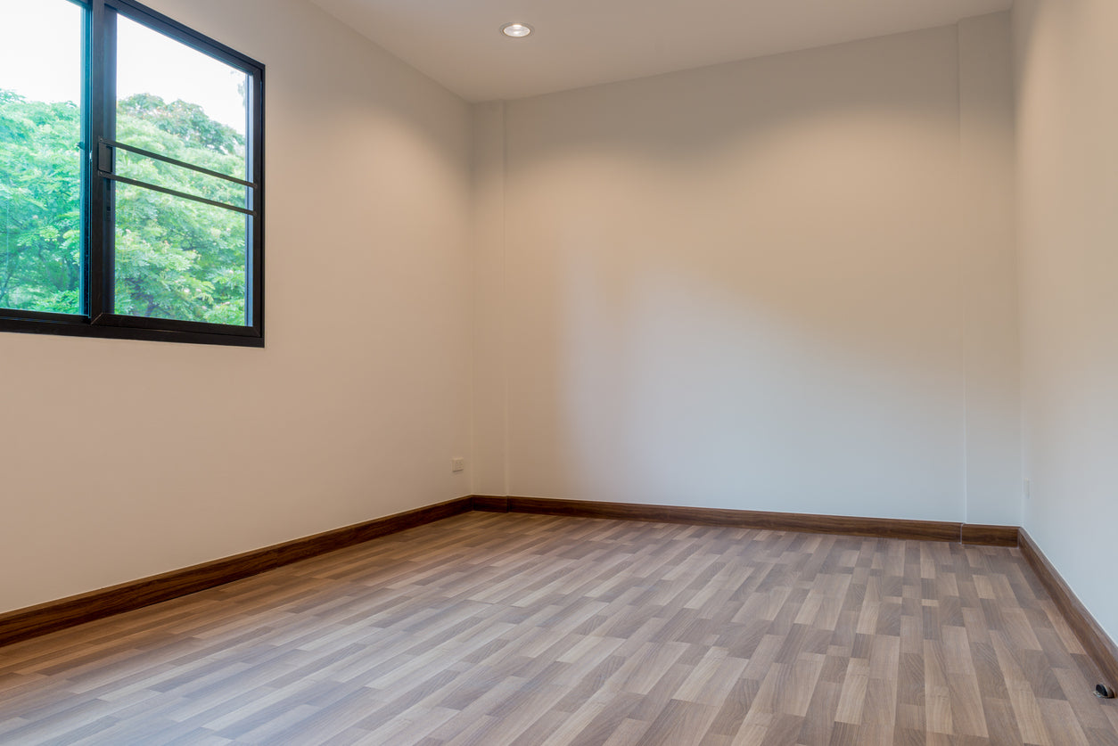 Chambre rénovée avec murs peints en beige clair, revêtement de sol stratifié aspect bois, plinthes assorties et fenêtre à cadre noir offrant une belle luminosité, réalisée par La Haute Peinture en Haute-Savoie.